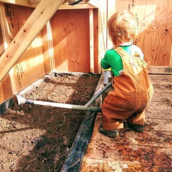 Toddler in overalls standing in a greenhouse, using a gardening tool to tend to a planting bed filled with soil. Sunlight filters through the wooden structure, highlighting the child's engagement in gardening activities.