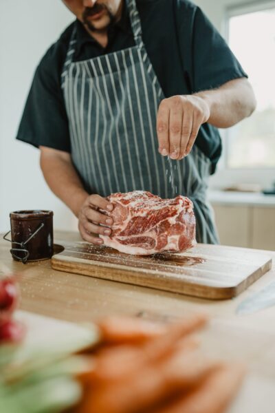 Person seasoning a raw pork roast with salt on a wooden cutting board in a kitchen setting, with fresh vegetables in the foreground and a jar nearby.